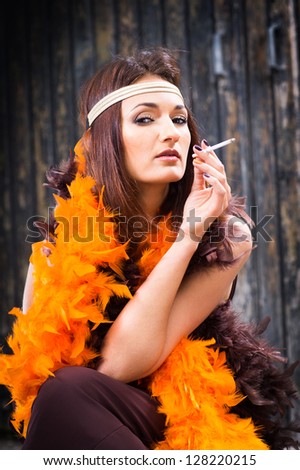 beautiful actress in brown and orange boa smoking against old wooden gate