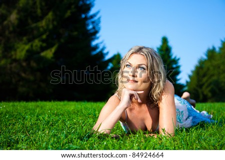 Portrait of attractive woman in blue sundress lying on green fresh grass and waiting for dawn against natural landscape at summer morning