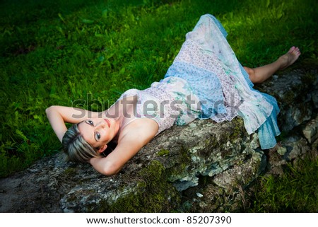 Portrait of attractive woman in blue sundress lying on stones against natural landscape at summer morning