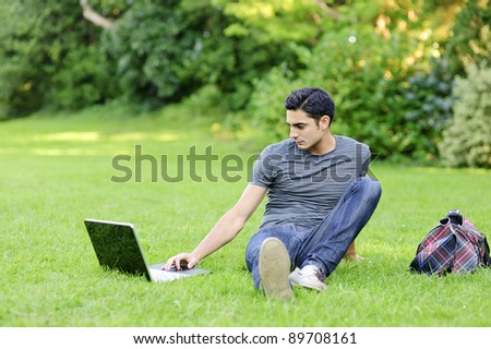 Handsome brunet young man in casual clothing sitting in the park on the grass and surf internet