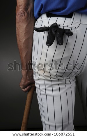 Closeup of a baseball player seen from behind and leaning on a wood bat. Man is unrecognizable with a batting glove in his pocket. Vertical format over a light to dark background.