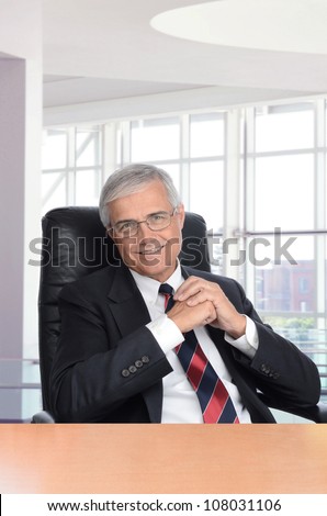 Smiling middle aged businessman with his hands together seated at a desk in a modern office building. Vertical format.