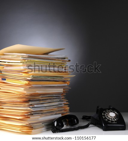 A large stack of files on a desk and a retro style telephone with the receiver off the hook. Square format over a light to dark gray background.