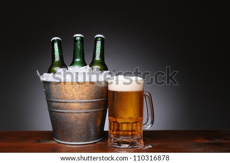 An ice bucket with three green beer bottles next to a full mug of ale on a wet wood surface. Horizontal format with a light to dark gray background.
