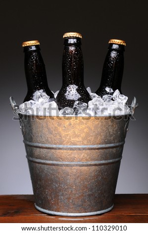 Ice bucket with three brown bottles of beer on a wet wood bar counter top. Vertical format on a light to dark gray background.