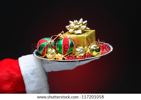 Santa Claus hand holding a tray with decorations and a gift. Hand and arm only on a light to dark red background.