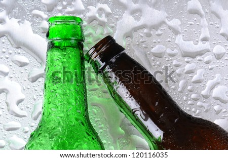 Closeup of two beer bottles on a wet stainless steel surface. One bottle is green the other brown. The brown bottle is at an angle. Overhead view.