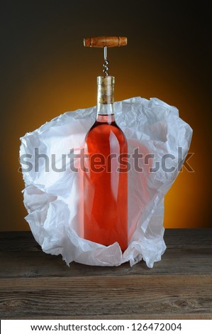 A bottle of blush wine wrapped in tissue paper, on a rustic wood surface and a light to dark warm background. A vintage cork screw is inserted in the bottle.