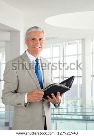 Portrait of a middle aged businessman standing in a modern office. Man is holding a small binder and smiling at the camera.