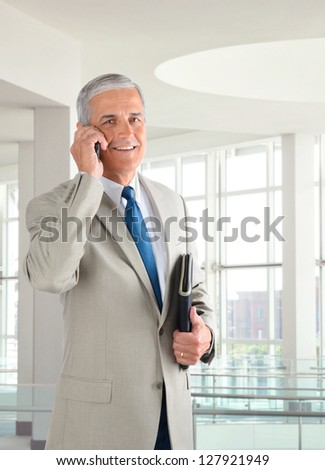 Portrait of a middle aged businessman standing in a modern office talking on a cell phone.. Man is holding a small binder and smiling at the camera.