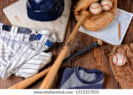 Overhead view of baseball gear on a rustic wood surface. Items include, Home Plate, helmet, baseball, ball, glove, catchers mitt, score book, pants, belt and bats.