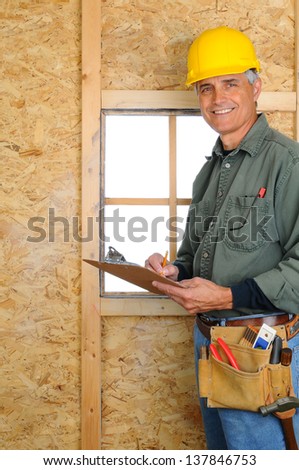 A middle aged contractor standing in new construction writing on a clip board. Man is wearing jeans, work shirt, tool belt and a hard hat, and smiling at the camera. Vertical Format.
