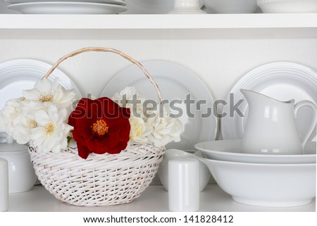 Closeup of a  basket of roses on the shelf of a cupboard full of white plates. Items include, plates, saucers, bowls and a gravy boat. There is one red rose amongst all the white.
