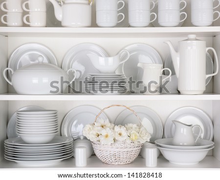 Closeup of white plates and dinnerware in a cupboard. A basket of white roses is centered on the bottom shelf. Items include, plates, coffee cups, saucers, soup tureen, tea pot, and gravy boats.