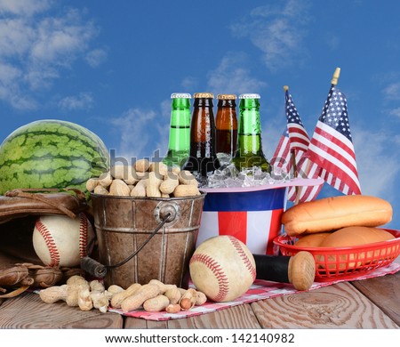 Picnic table ready for a Fourth of July celebration. Cold beer in an Uncle Sam Hat, watermelon peanuts, baseball, and hot dog buns fill the table, with a blue cloudy sky background.