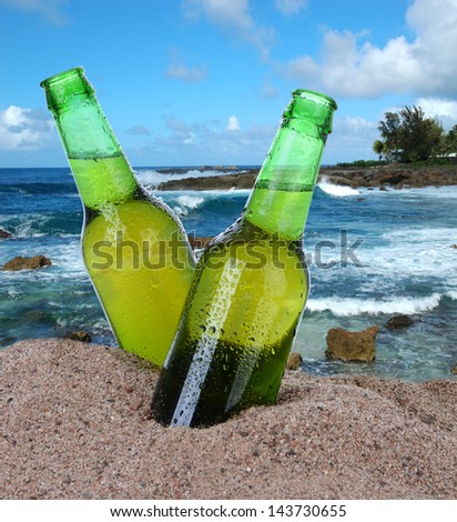 Closeup of two beer bottles in the sand with a tropical ocean background. Square format. Bottles are covered with condensation.