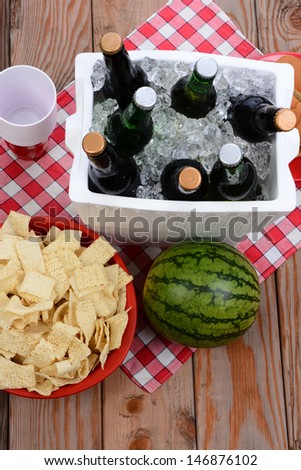 High angle shot of a picnic spread on a wood deck with red and white checkered table cloth. Items include: beer, ice chest, cups, chips and watermelon