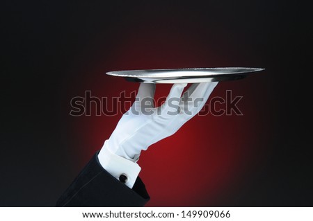 Closeup of a waiter\'s hand holding a silver serving tray in his fingertips over a light ot dark red background. Only the man\'s hand and arm are visible. He is wearing a tuxedo and formal white gloves.