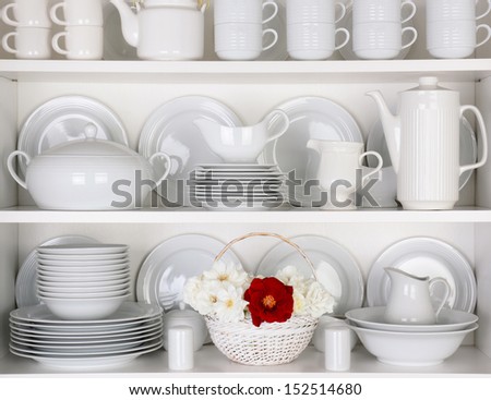 Closeup of white plates and dinnerware in a cupboard. A basket of white and red roses is centered on the bottom shelf. Items include, plates, coffee cups, saucers, soup tureen, tea pot, and gray boats