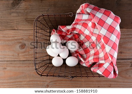 A wire basket with eggs and a red checkered napkin on a rustic wood surface. Overhead shot looking straight down into the basket.