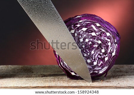 A head of red cabbage and a carving knife. Horizontal format on a rustic wood surface and a warm light to dark background. The cabbage and knife are covered with water droplets.