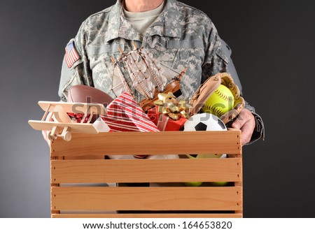 Closeup of a soldier in fatigues holding a wooden box full of toys and sports equipment for a holiday charity drive. Horizontal format man is unrecognizable.