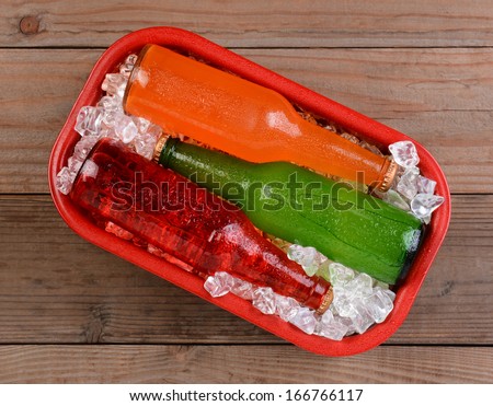Overhead shot of sodas on ice in a red metal cooler. Three different drinks, strawberry , orange, and lemon lime. The cooler is sitting on a rustic wooden surface.
