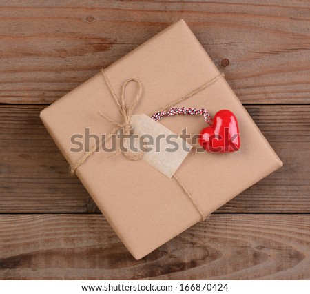 High angle view of a plain brown paper wrapped Valentines Day present with a ceramic red heart. Square format on a rustic wood surface.
