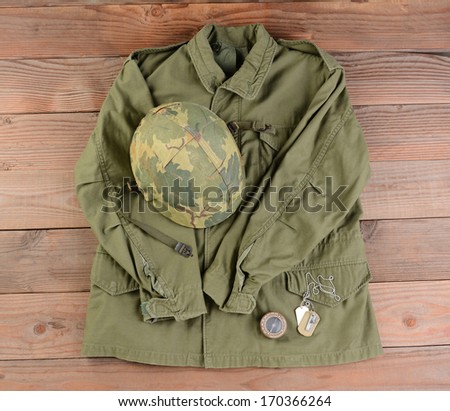 High angle view of an Army Field Jacket and Helmet on a rustic wood floor. Other items include dog tags and Army Compass. The vintage gear is from the Vietnam Era.
