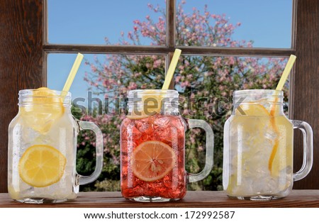 Glasses of lemonade and fruit Juice on a window ledge on a bright sunny summer day. The mason jar style glasses have handles and drinking straws. Thru the window is a tree and blue sky.