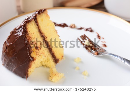 A slice of yellow cake with chocolate frosting with a piece missing. The white plate and fork have crumbs and frosting.
