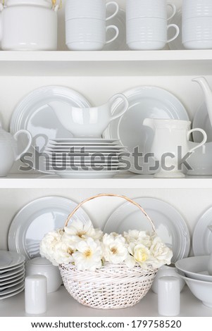 Vertical closeup of a  basket of roses on the shelf of a cupboard full of white plates. Items include, plates, saucers, bowls and a gravy boat.