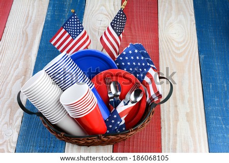 High angle photo of a picnic table ready for Fourth of July Party. A Basket full of Cups, plates, utensils, napkins and American Flags sits in the middle of the red white and blue table.