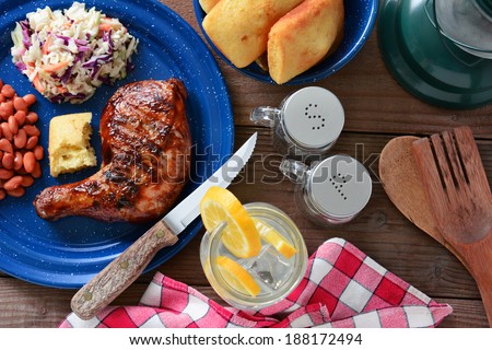 High angle shot of a barbecue chicken plate with cole slaw, pinto beans and corn bread. The meal is on a rustic wooden table with a red and white checked napkin and a glass of lemonade.