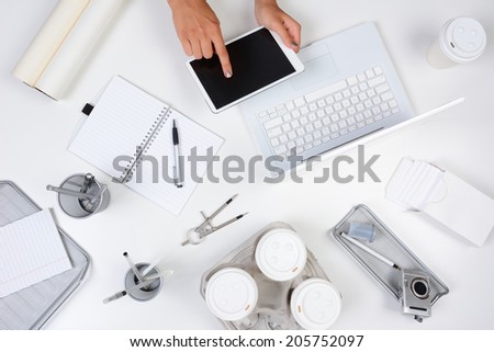 High angle shot of a white desk with primarily white and silver office objects, with a woman holding a tablet computer showing her hands only. Tablet has a blank screen.