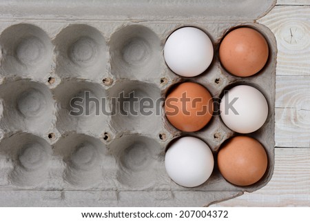 Six Eggs, three white and three brown, in a large carton with empty spaces. The carton is on a white farmhouse style wood kitchen table.
