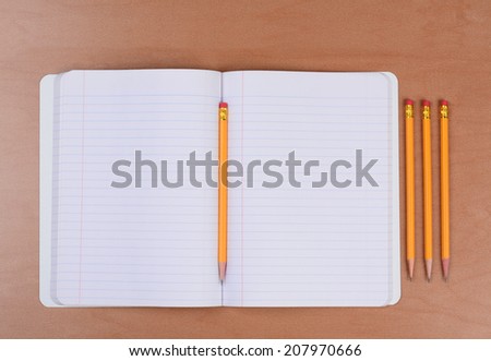 An open theme book with a pencil in the fold and three more sharpened pencils to the side. Horizontal format on a wood classroom desk.