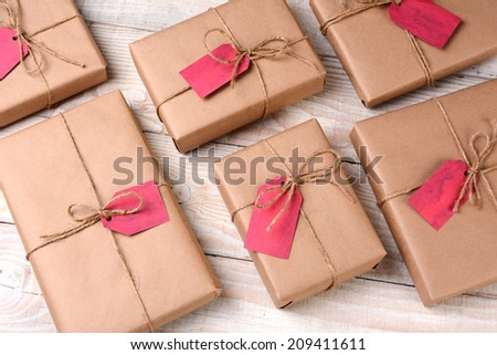 A group of plain brown paper wrapped Christmas presents on a white wood table. The gifts are tied with twine and have blank red gift tags. High angle shot.