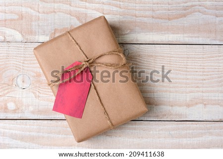 High angle shot of a plain brown paper wrapped Christmas present on a whitewashed wood table. The gift is tied with twine with a blank red gift tag.