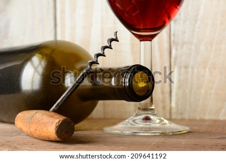 Wine still life with a bottle on its side and the bottom of a glass of red wine and a cork screw leaning on the bottle.  Horizontal format with shallow depth of field on a wood background.
