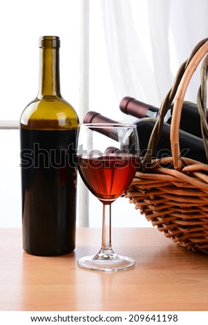 Wine still life with a basket of bottles and a glass of red wine in front of a window with curtains. Vertical format.