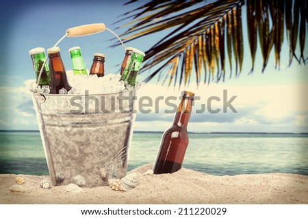 Assorted beer bottles in a bucket of ice in the sand on a tropical beach with an instagram look. One beer bottle without a cap is by itself stuck in the sand next to the pail.