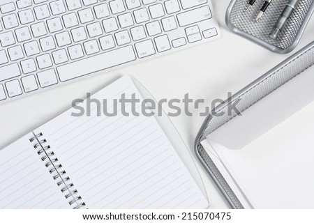 High angle shot of a white desk with primarily white and silver office objects. Items include, Keyboard, pad, pens, compass, and In-box tray. Horizontal format with copy space.