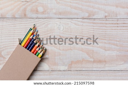 High angle shot of a box of multi-colored pencils on a white wood table. The pencils are partially out of the box and at an angle in the bottom left corner, with copy space.