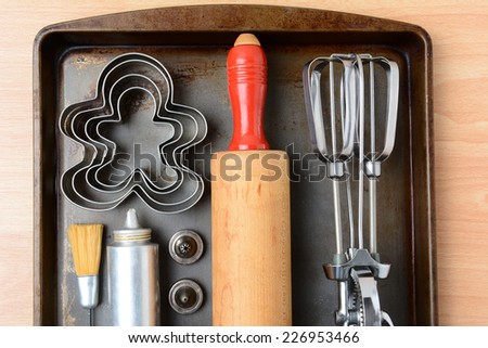 High angle closeup of the kitchen tools needed for making holiday cookies. The items are arranged on a well used metal baking sheet. Horizontal format with rolling pin, mixer, cookie cutters and press