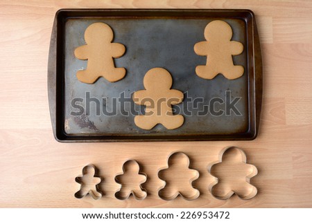 High angle shot of a cookie sheet with three gingerbread man cookies. Below the pan is a row of four cookie cutters of different sizes, Horizontal format on a butcher block kitchen table.