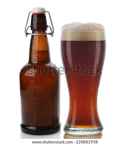 Closeup of a glass of dark beer with a frothy head next to a swing top brown beer bottle. Straight on shot on a white background with reflection. Both items are cover with water drops.
