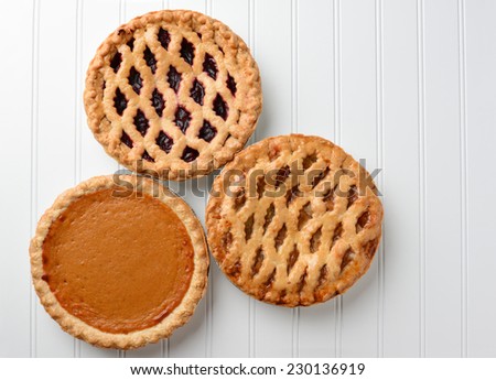 High angle shot of three pies, apple, pumpkin, and cherry. Horizontal format on a white beadboard surface. The pies are favorites for the  American Holidays, Thanksgiving and Christmas.