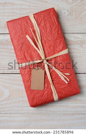 High angle image of a red tissue wrapped Christmas present on a rustic white wood kitchen table. The gift is tied with raffia and has a blank gift tag. Vertical Format.