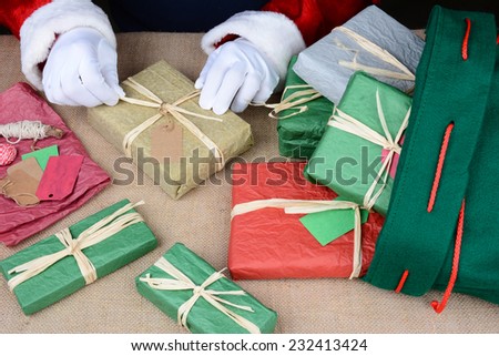 Closeup of Santa Claus wrapping presents. Only Santa\'s hands are visible as he puts the finishing touches on a variety of gift boxes next to his bag on Christmas Eve. Horizontal Format.